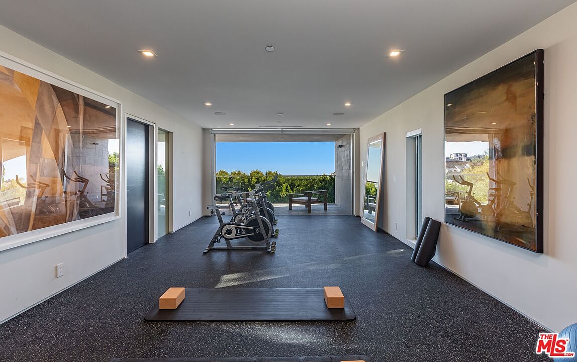 This modern home gym features dark, textured flooring and a minimalist aesthetic, centered around multiple stationary exercise bikes. Large, abstract art pieces adorn the side walls, while a yoga mat with two foam blocks sits in the foreground. The room opens directly onto a covered outdoor deck through a wide, unobstructed opening, providing a seamless transition to the scenic hillside view beyond.