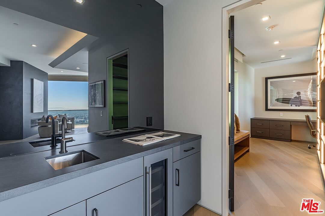 This image captures a sleek, modern wet bar area featuring dark cabinetry, a stainless steel sink, and a built-in wine refrigerator. A doorway leads into a bright, adjacent office space furnished with a desk and a large framed artwork on the wall. The perspective highlights the clean lines and contemporary aesthetic of the home, with a reflective dark wall panel adding depth to the space.