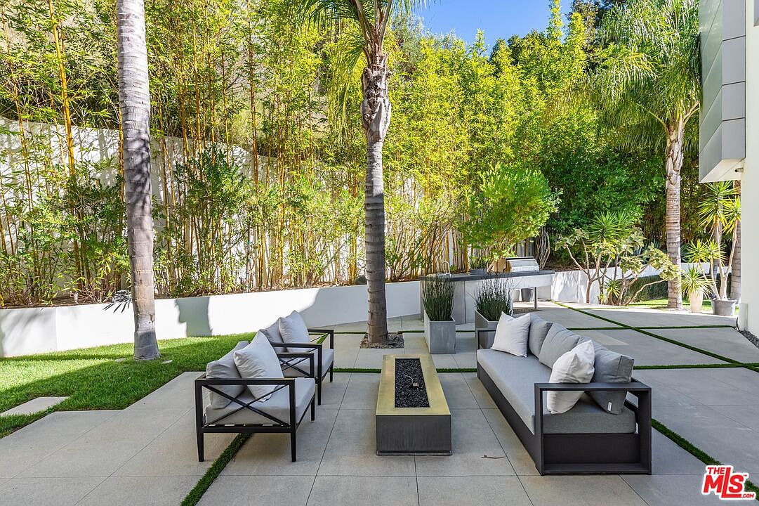 This modern outdoor patio features a sleek, rectangular fire pit centered between a contemporary sofa and two matching armchairs with light-colored cushions. The space is paved with large concrete tiles and is framed by lush bamboo landscaping and tall palm trees, creating a private, resort-like atmosphere. In the background, an outdoor kitchen area with a grill is visible, enhancing the space's suitability for al fresco dining and entertaining.