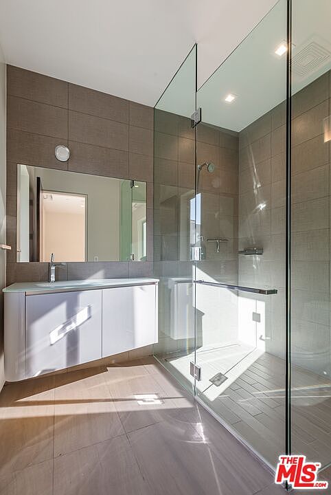 This modern bathroom features a sleek, wall-mounted white vanity with a single sink and a large, frameless mirror. Adjacent to the vanity is a spacious walk-in shower enclosed by floor-to-ceiling glass panels, showcasing neutral-toned wall tiles and a matching tiled floor. The space is illuminated by recessed lighting, creating a clean, minimalist, and contemporary aesthetic.