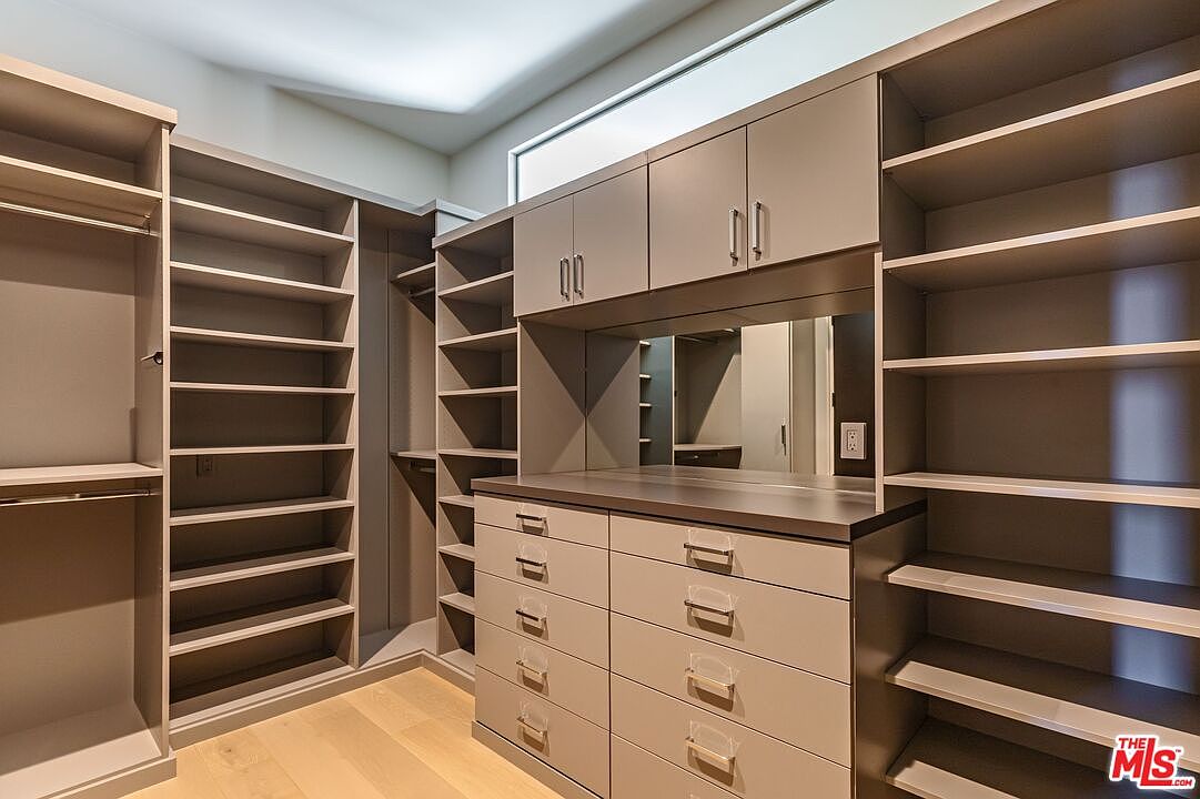 This spacious walk-in closet features custom light-toned cabinetry with a mix of open shelving, hanging rods, and a central bank of drawers. A built-in mirror above the counter adds depth to the room, while a transom window above the cabinets allows for natural light. The clean, organized layout and neutral color palette create a sophisticated and functional storage space.