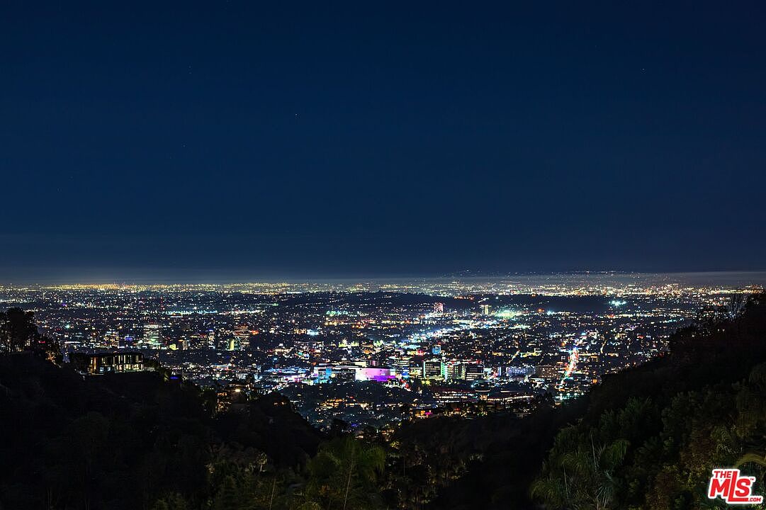 This image captures a breathtaking, wide-angle night view of a sprawling city skyline from an elevated vantage point. The scene is dominated by a dense grid of glowing urban lights that stretch toward the horizon, framed by dark, silhouetted hills in the foreground. The perspective suggests a high-end property location with a dramatic, panoramic outlook over the metropolitan area.