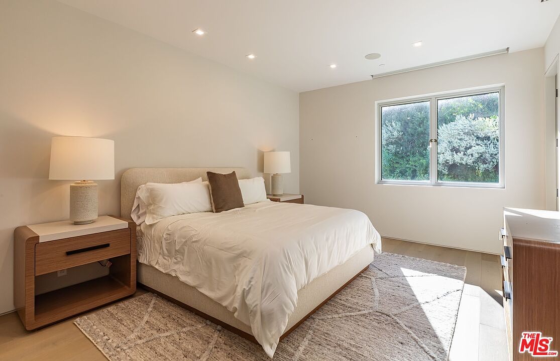 This bright and airy primary bedroom features a neutral color palette with a plush, upholstered bed as the central focal point. Two matching light wood nightstands with modern lamps flank the bed, which is dressed in crisp white linens and accented by a single dark throw pillow. A large window allows natural light to fill the room, highlighting the textured area rug and the clean, minimalist aesthetic of the space.