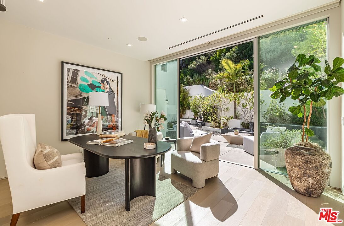This modern home office features a sleek, dark-toned desk paired with a white high-back chair, set against a backdrop of light-toned walls and light wood flooring. A large piece of abstract art hangs above the desk, while a large potted plant adds a touch of greenery near the expansive glass sliding doors. The room opens directly onto a sunlit patio, creating a seamless transition between the indoor workspace and the outdoor garden area.
