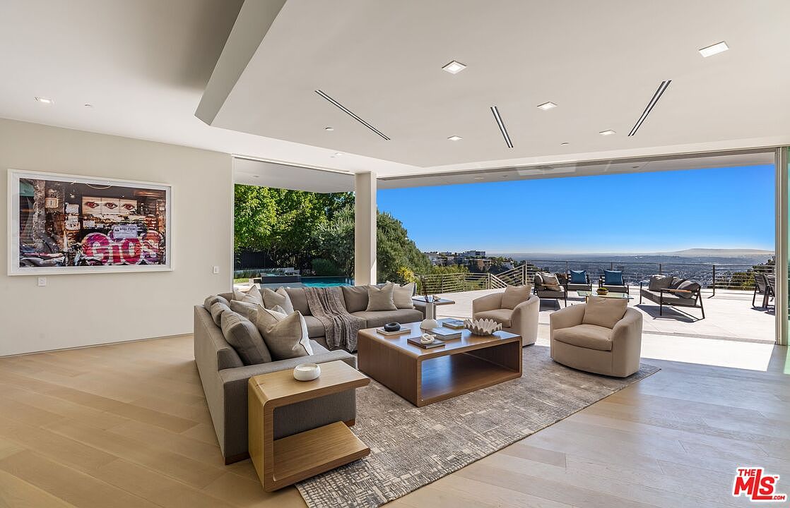 This expansive, open-concept living room features a neutral color palette and light wood flooring, centered around a large sectional sofa and a low-profile wooden coffee table. The space seamlessly transitions to an outdoor terrace through massive, retractable glass walls that offer panoramic views of the city and hills beyond. A large, vibrant piece of wall art adds a pop of color to the minimalist interior, while recessed lighting and a clean, modern aesthetic define the sophisticated atmosphere.