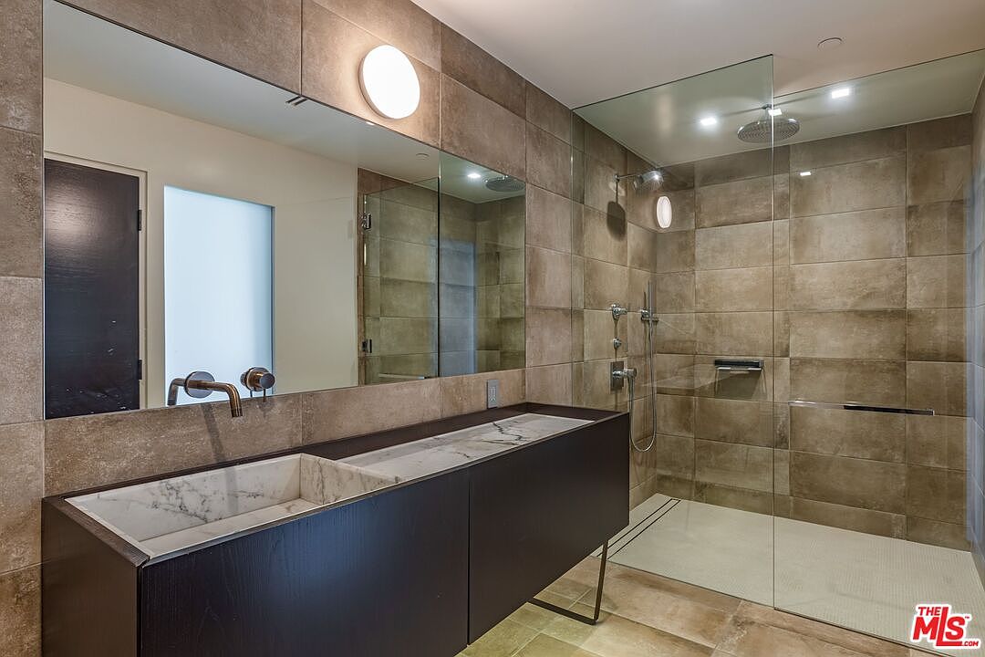 This modern bathroom features a sleek, dark-toned vanity with a stone-patterned integrated sink and a wall-mounted faucet. A large, frameless glass-enclosed walk-in shower with neutral-toned wall tiles and a rainfall showerhead occupies the right side of the space. The room is illuminated by a circular wall-mounted light fixture and recessed ceiling lights, creating a clean and sophisticated aesthetic.