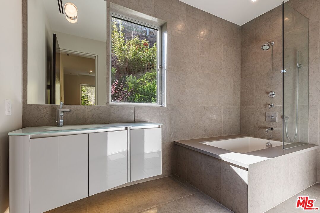 This modern bathroom features a sleek, light-toned vanity with a glass countertop and a large mirror reflecting the space. A deep soaking tub is integrated into a tiled platform, which extends into a walk-in shower area enclosed by a glass panel. The room is finished with neutral-toned wall tiles and a window that provides natural light and a view of the outdoors.