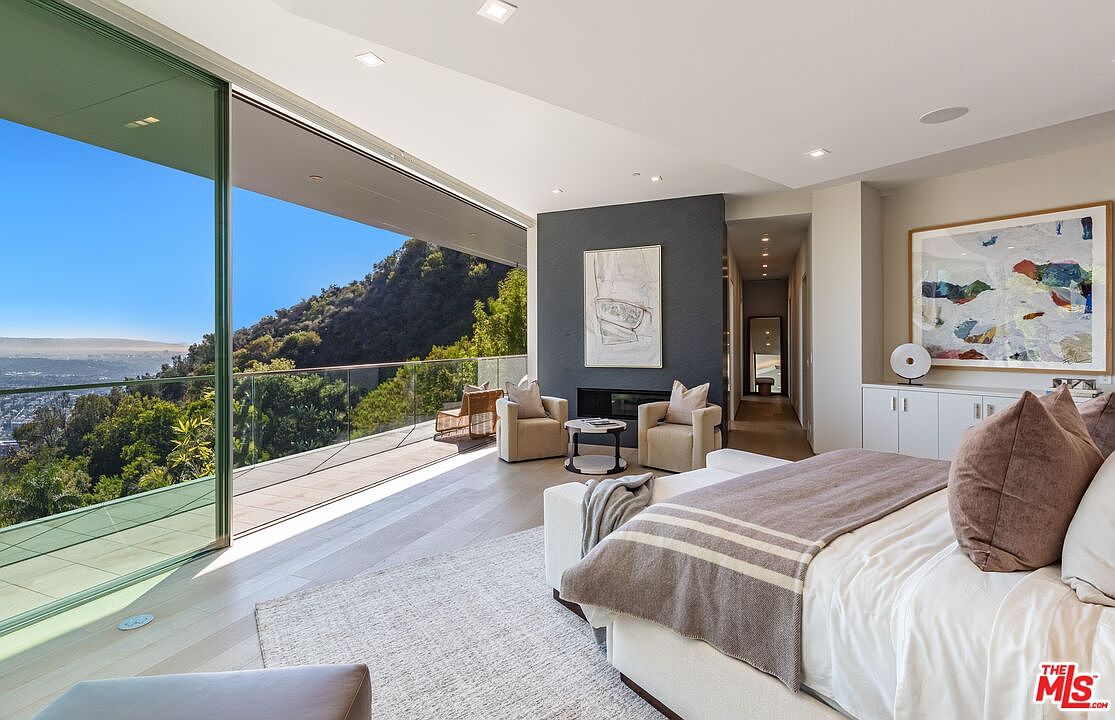 This luxurious primary bedroom features a minimalist aesthetic with a neutral color palette and expansive floor-to-ceiling glass walls that provide panoramic views of the surrounding hills. The room includes a cozy seating area with two armchairs centered around a dark, textured chimney breast, and a plush bed dressed in soft linens. The space flows seamlessly onto a private balcony, creating an airy, indoor-outdoor living experience.