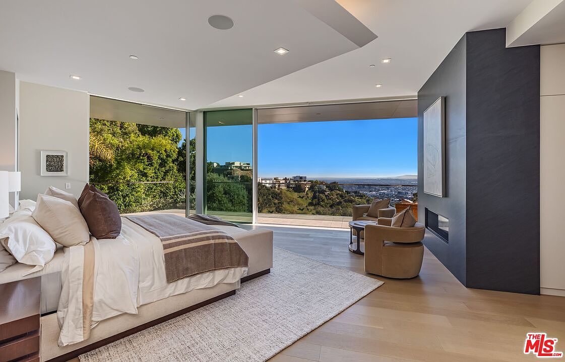 This spacious primary bedroom features a contemporary design with light wood flooring and a neutral color palette. A large bed with plush bedding sits center stage, while floor-to-ceiling glass walls provide an expansive, unobstructed view of the surrounding hills and city skyline. To the right, a dark, full-height surround fireplace adds a dramatic focal point, complemented by a cozy seating area with two armchairs.