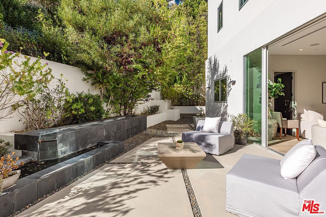 This modern outdoor patio space features a sleek, dark-tiled water feature and a minimalist seating area with light-colored lounge chairs. The area transitions seamlessly into the interior living space through large sliding glass doors, creating an ideal indoor-outdoor flow. Lush greenery and a stone-lined path provide a serene, private atmosphere for this contemporary backyard retreat.