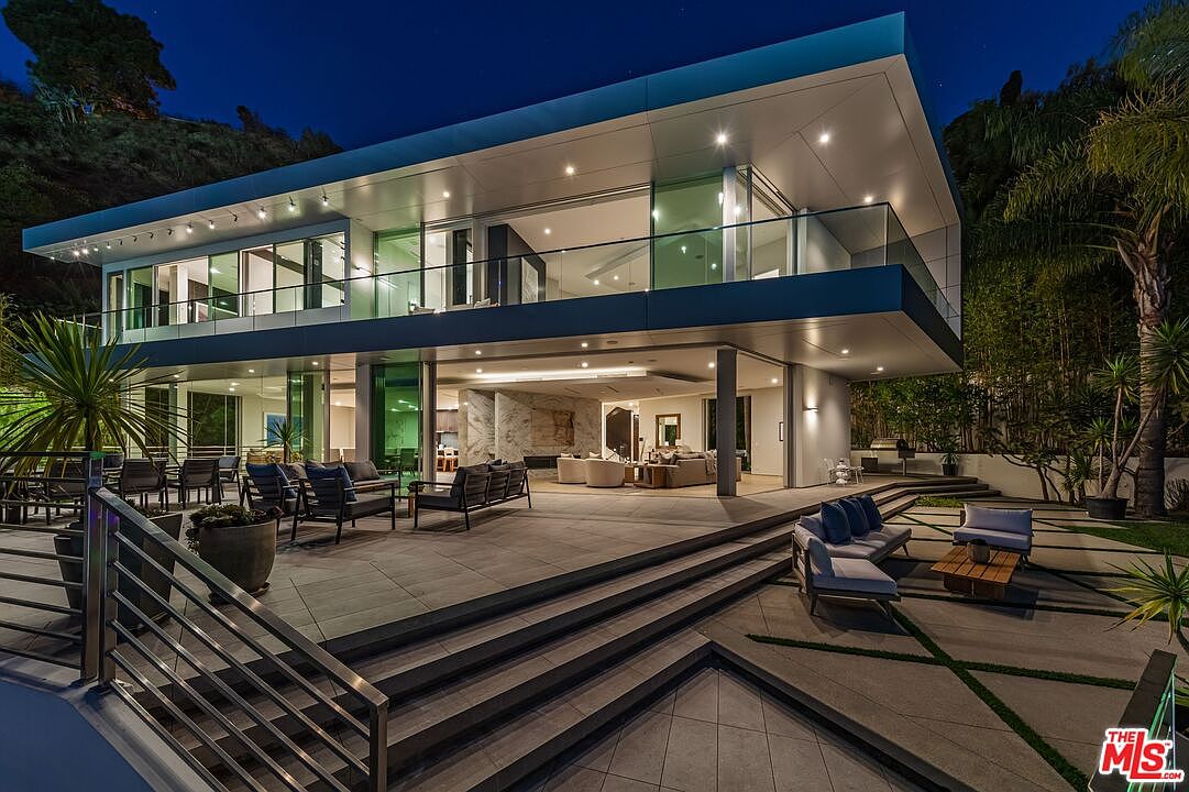 This contemporary luxury residence features a striking architectural design with a cantilevered upper level and extensive glass walls that create a seamless indoor-outdoor flow. The expansive stone patio includes multiple seating areas, tiered steps, and integrated lighting, all set against a backdrop of lush hillside landscaping. The evening lighting highlights the clean lines and modern aesthetic of the home, emphasizing its sophisticated and open-concept design.