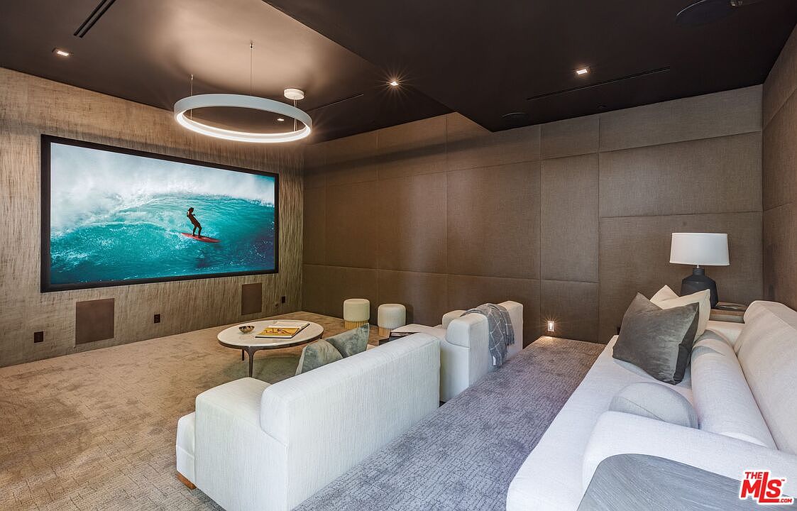 This sophisticated home theater features a large projection screen displaying a surfing scene, set against a wall with textured, paneled acoustic treatments. The room is furnished with plush, tiered seating and a central circular coffee table, all illuminated by a modern, circular pendant light fixture. The dark ceiling and neutral-toned walls create an immersive, cinematic atmosphere perfect for entertainment.