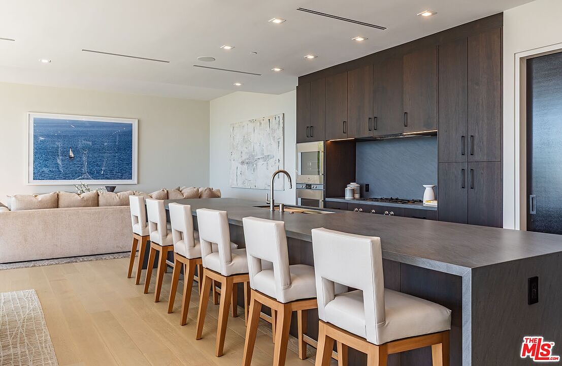 This modern, open-concept kitchen features a large, dark-toned island with integrated seating for five, serving as the focal point of the space. Behind the island, dark cabinetry provides a sleek contrast against the light-colored walls and light wood flooring. The area flows seamlessly into a living space adorned with a large coastal-themed artwork, creating a bright and airy atmosphere suitable for contemporary living.
