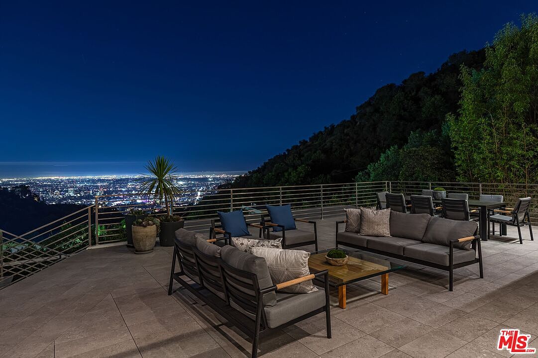 This expansive outdoor terrace offers a panoramic view of a sprawling city skyline at night, set against a backdrop of dark, forested hills. The space is furnished with contemporary dark-toned patio furniture, including a sofa, armchairs, and a dining set, all arranged on a neutral-toned tiled surface. A metal railing borders the edge of the deck, emphasizing the elevated perspective and the dramatic, open-air atmosphere.