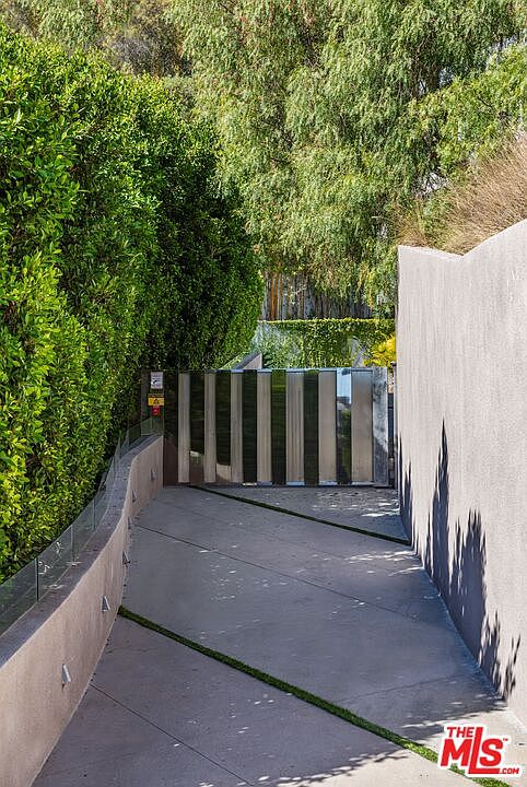 This image captures a private, gated driveway leading toward a modern residence, framed by tall, manicured hedges on the left and a smooth, light-colored concrete wall on the right. The driveway features a geometric design with concrete sections separated by thin strips of grass, leading to a sleek, vertical-slat metal gate. The perspective is from the bottom of the incline, looking upward toward the entrance, creating a sense of exclusivity and architectural privacy.