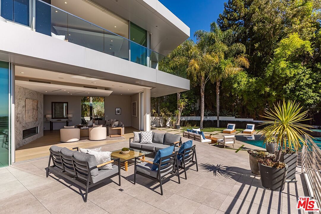 This modern luxury residence features a seamless indoor-outdoor transition with a spacious stone-paved patio overlooking a private swimming pool. The outdoor living area is furnished with contemporary lounge seating, while the interior living space is visible through expansive glass walls, showcasing a minimalist design with a stone-clad fireplace. The scene is framed by lush greenery and tall palm trees, creating a serene and sophisticated atmosphere perfect for entertaining.