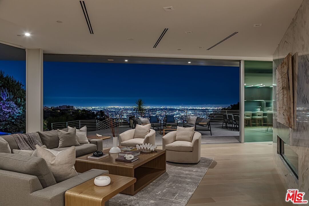 This sophisticated living room features an open-concept design that seamlessly transitions to an expansive outdoor deck through large glass panels. The interior is furnished with plush, neutral-toned seating arranged around a central coffee table, while a tall stone-clad chimney breast anchors the side wall. The space offers a breathtaking, panoramic view of the city lights at night, creating a luxurious and serene atmosphere.