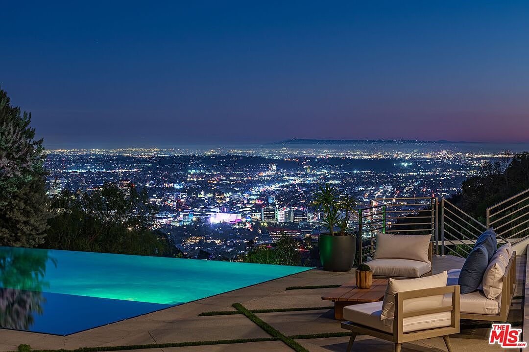 This stunning outdoor terrace features an infinity-edge pool that seamlessly blends into the expansive, panoramic night view of the city lights below. The patio is furnished with comfortable lounge seating, creating an ideal space for evening relaxation and entertaining. The cinematic perspective captures the serene transition from the illuminated pool water to the vast, glowing urban horizon under a twilight sky.