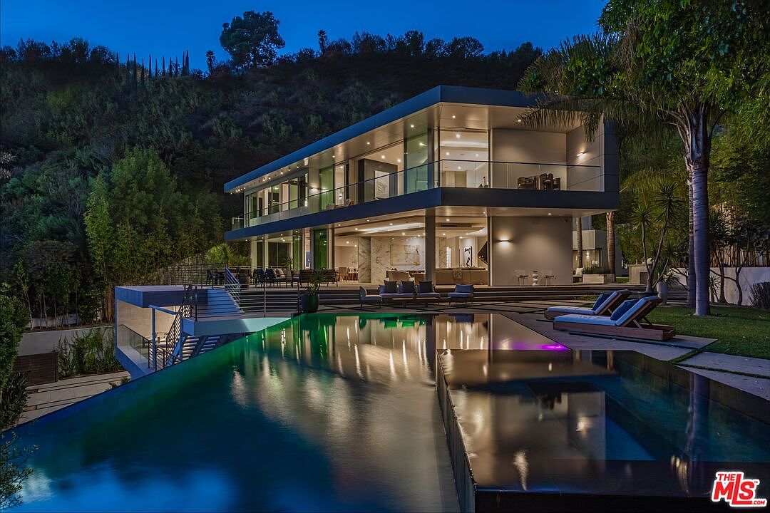 This stunning contemporary residence is captured at twilight, showcasing a multi-level architectural design with extensive glass walls that blur the line between indoor and outdoor living. A sleek, dark-bottom infinity pool dominates the foreground, reflecting the warm interior lighting and the surrounding hillside landscape. The expansive patio area features comfortable lounge seating, creating a sophisticated atmosphere perfect for evening entertaining.