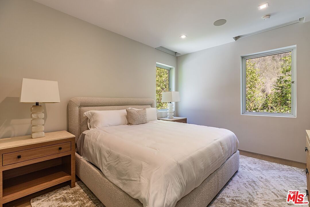 This bright and serene bedroom features a plush, upholstered bed frame centered against a neutral-toned wall. Two light wood nightstands with matching table lamps flank the bed, while a patterned area rug adds texture to the space. Large windows allow natural light to fill the room, offering a view of the hillside greenery outside.