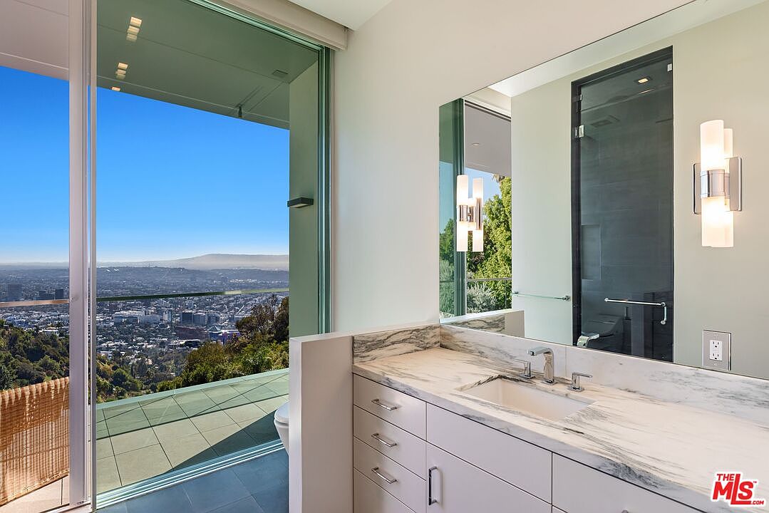 This modern bathroom features a sleek vanity with a stone countertop and integrated sink, complemented by a large mirror that reflects the expansive city view. Floor-to-ceiling glass sliding doors open onto a private balcony, seamlessly blending the indoor space with the outdoors. The minimalist design and neutral color palette create a sophisticated and airy atmosphere.