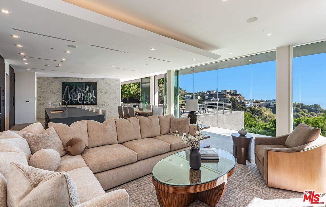 This expansive, open-concept living area features a large, plush sectional sofa and a central oval coffee table, creating a comfortable space for relaxation. The room is defined by its seamless connection to the outdoors through expansive glass walls that offer panoramic views of the surrounding hills. In the background, a sleek kitchen island with a dark surface sits against a wall with a prominent stone-clad feature, while a dining area is visible further back, all illuminated by recessed lighting and natural sunlight.
