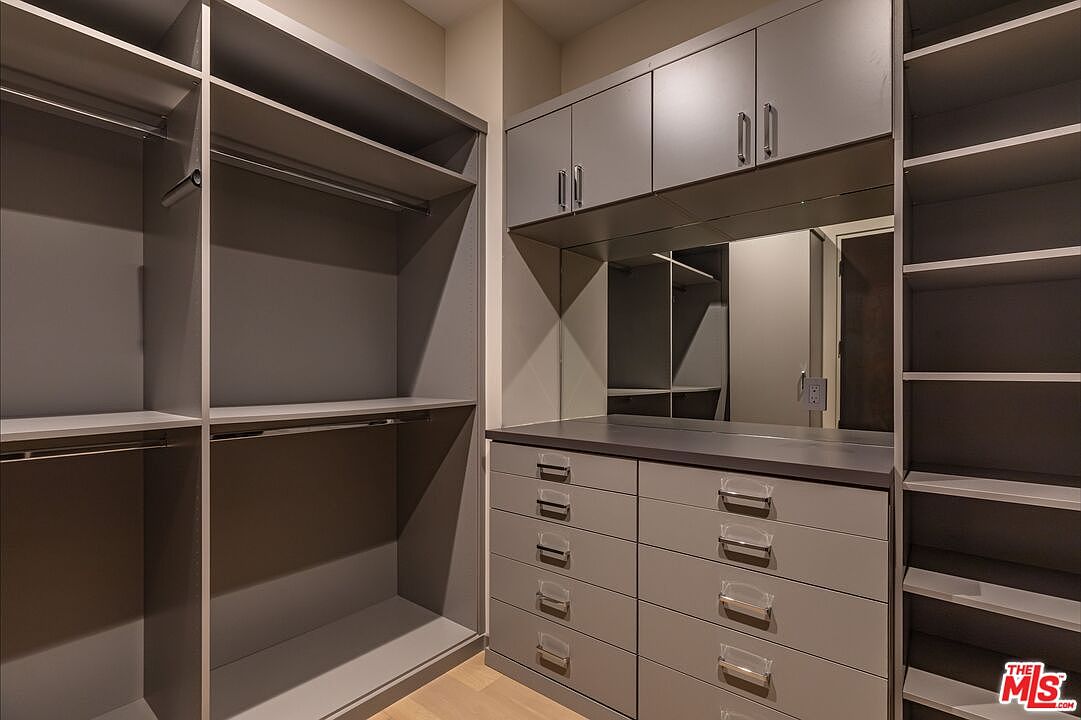 This organized walk-in closet features custom grey cabinetry with a mix of open shelving, hanging rods, and a bank of drawers. A central vanity area includes a mirror and a flat surface, providing a functional space for dressing. The neutral color palette and clean lines create a modern and sophisticated storage solution.
