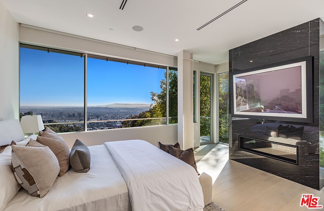 This modern primary bedroom features a large bed positioned to take advantage of expansive, panoramic city views through floor-to-ceiling windows. A striking black stone-clad chimney breast serves as a focal point, housing a built-in television and a linear fireplace below. The room is finished with light wood flooring and a neutral color palette, creating a sophisticated and serene atmosphere.