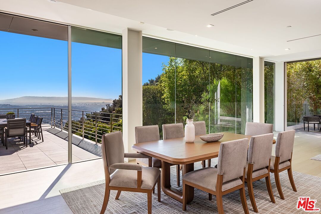 This modern dining room features a large wooden table surrounded by eight upholstered chairs, set atop a textured area rug. The space is defined by expansive floor-to-ceiling glass walls that provide an unobstructed view of the outdoor deck and the scenic hillside landscape beyond. The minimalist design emphasizes an open, airy atmosphere, seamlessly blending the indoor dining area with the natural environment.