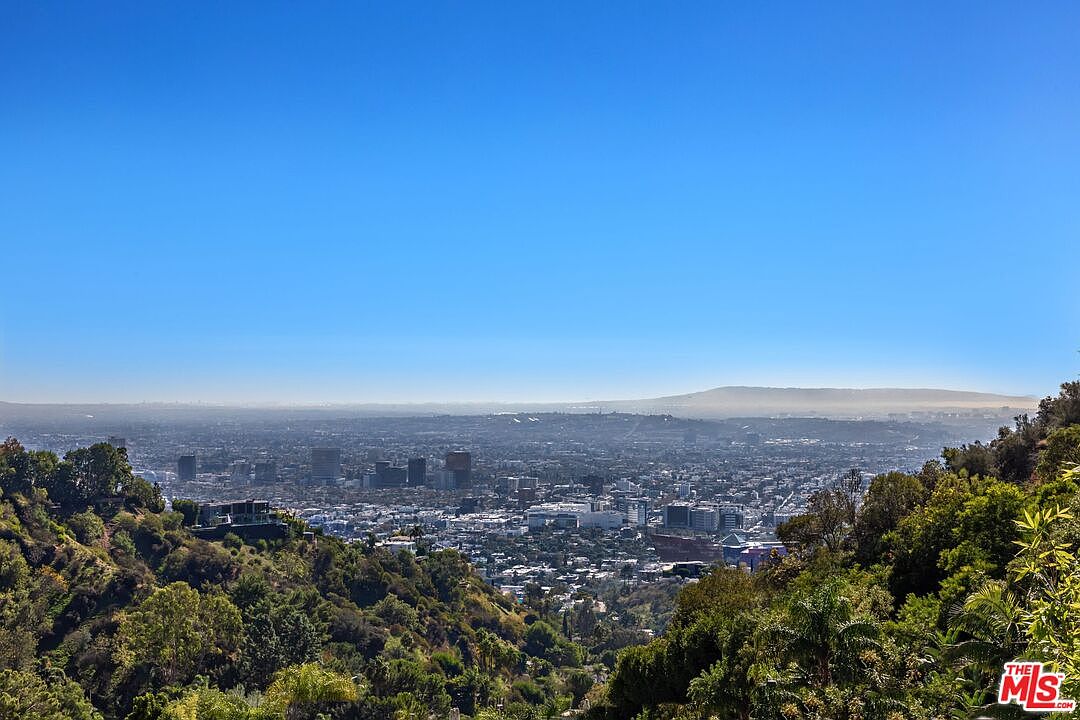 This image captures a sweeping, elevated panoramic view of a sprawling cityscape under a clear, bright blue sky. In the foreground, lush green hillsides frame the scene, with a modern residential structure partially visible on the left slope. The perspective is cinematic, looking out over the dense urban sprawl toward a distant, hazy mountain range on the horizon.