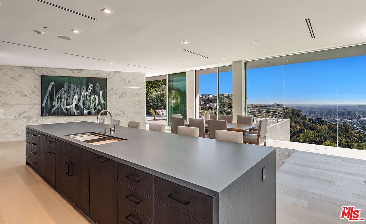 This modern, open-concept kitchen features a large, dark-toned central island with a built-in sink and ample storage. The space is defined by a striking stone-clad wall adorned with a large abstract painting, while expansive glass walls provide an unobstructed, panoramic view of the city skyline and surrounding hills. A dining area is seamlessly integrated into the open floor plan, creating a sophisticated and airy atmosphere perfect for entertaining.