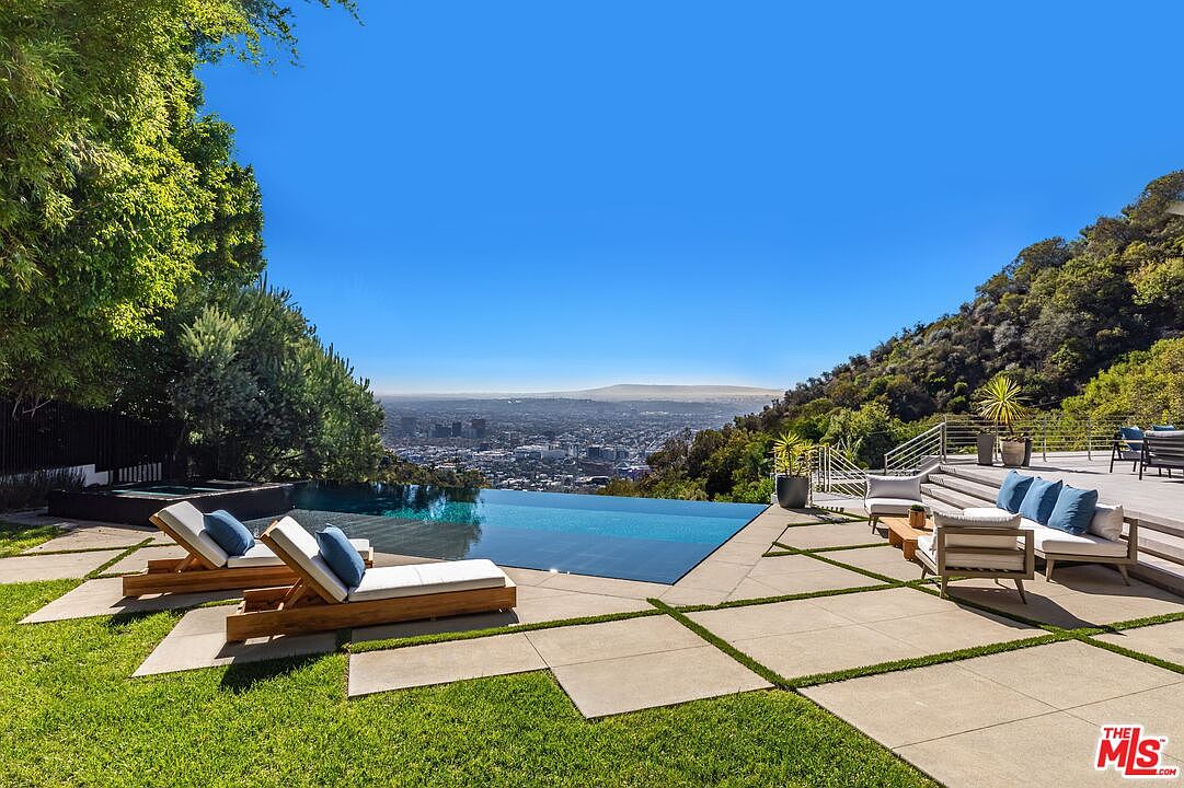 This stunning outdoor space features a sleek infinity-edge pool that seamlessly blends with the expansive panoramic city view in the background. The surrounding patio is composed of large, light-colored stone pavers set in a grid pattern, complemented by comfortable lounge chairs and outdoor seating. Lush green grass and mature trees frame the scene, creating a private and luxurious atmosphere perfect for entertaining.
