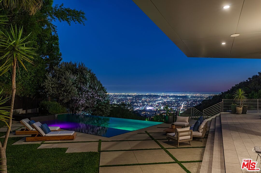 This stunning outdoor living space features a sleek infinity-edge pool that appears to merge with the expansive, glittering city lights below. The patio is paved with large, light-toned stone tiles and includes comfortable lounge seating, creating a sophisticated environment for evening relaxation. The perspective is captured from under a modern, covered overhang, emphasizing the seamless transition between the luxurious home and the dramatic panoramic view.
