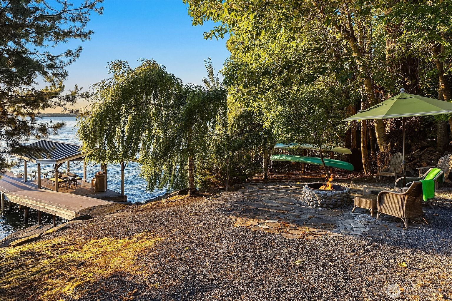 This serene waterfront outdoor space features a stone fire pit area nestled among mature trees, providing a perfect spot for relaxation by the lake. A wooden dock with a covered seating area extends into the water, while a green umbrella and wicker chairs offer comfortable lakeside lounging. The scene captures a peaceful, rustic atmosphere ideal for evening gatherings and enjoying the natural surroundings.