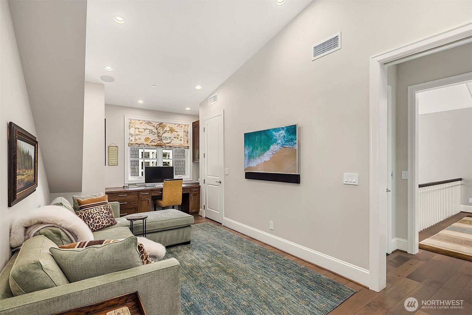 This versatile room functions as a cozy home office and lounge area, featuring a built-in wooden desk workspace positioned under a window with patterned roman shades. A comfortable green sectional sofa with decorative pillows and a textured area rug creates a relaxing seating area, while a wall-mounted television adds entertainment value. The space is bright and airy with high ceilings, neutral wall tones, and a seamless transition into the adjacent hallway.
