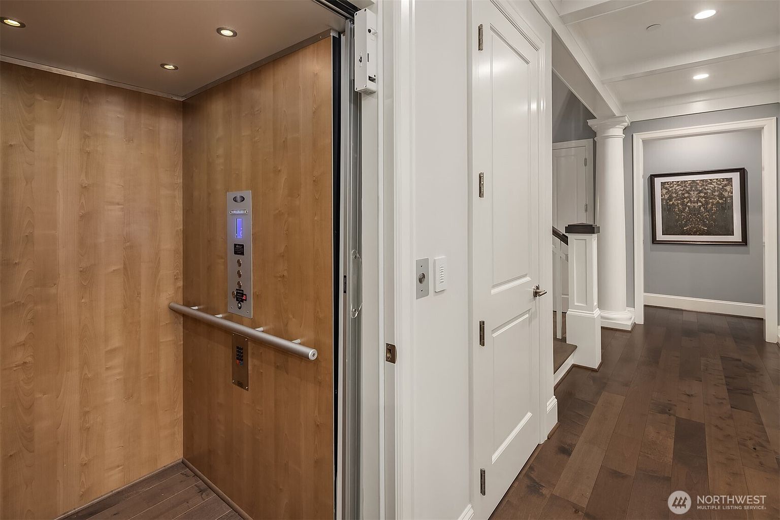 This image captures a residential elevator with a warm wood-paneled interior, positioned adjacent to a white paneled door and a hallway. The perspective looks into the elevator car, revealing a sleek control panel and handrail, while the adjacent hallway features dark hardwood floors, a classic framed artwork, and architectural columns. The space conveys a sense of luxury, accessibility, and refined traditional design.