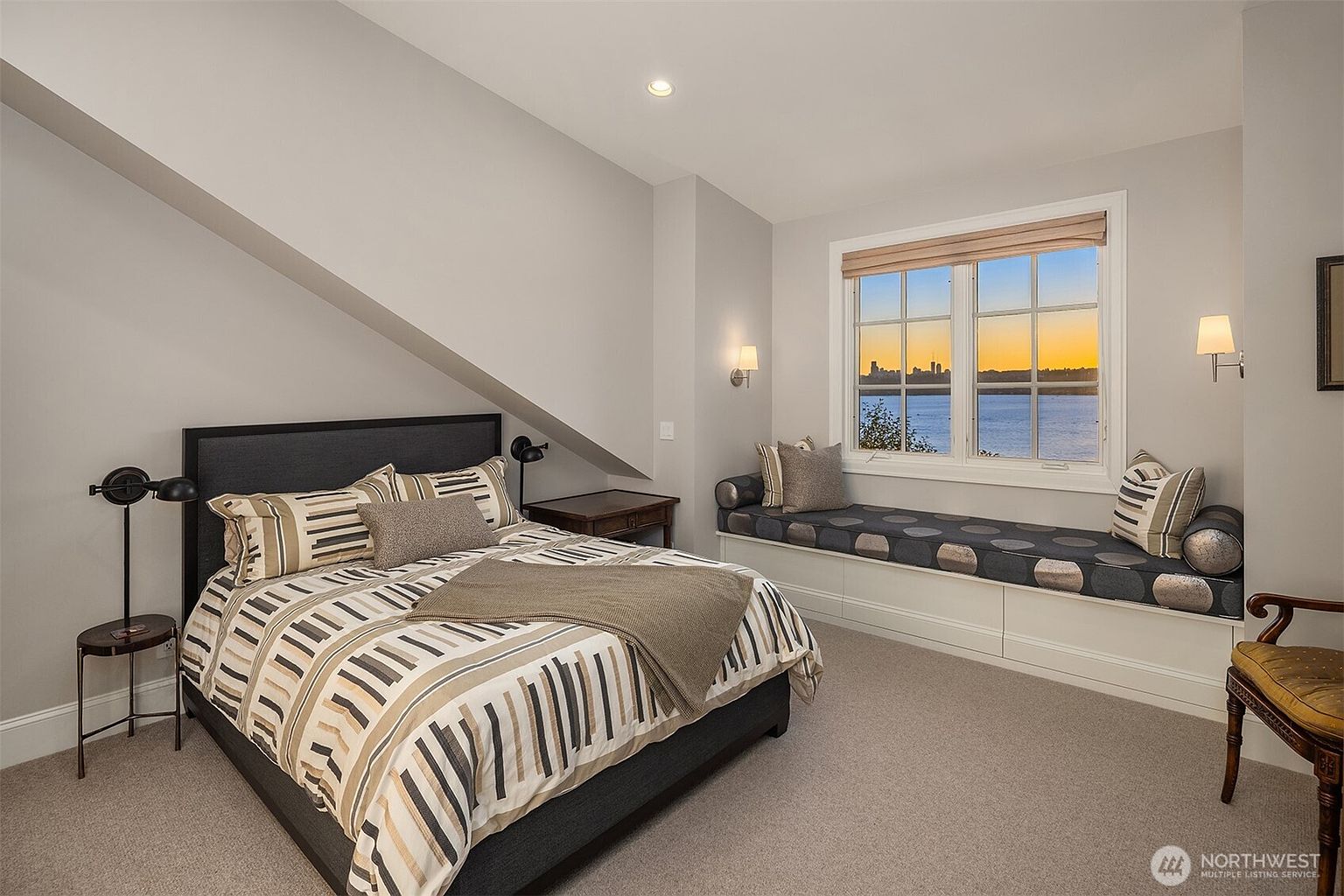 This cozy bedroom features a sloped ceiling, a queen-sized bed with a striped comforter, and a built-in window seat offering a scenic view of the water and city skyline at sunset. The room is decorated in a neutral palette with warm lighting from wall sconces and a bedside lamp, creating a tranquil and inviting atmosphere. The perspective is a wide-angle shot from the corner of the room, capturing both the sleeping area and the comfortable seating nook.
