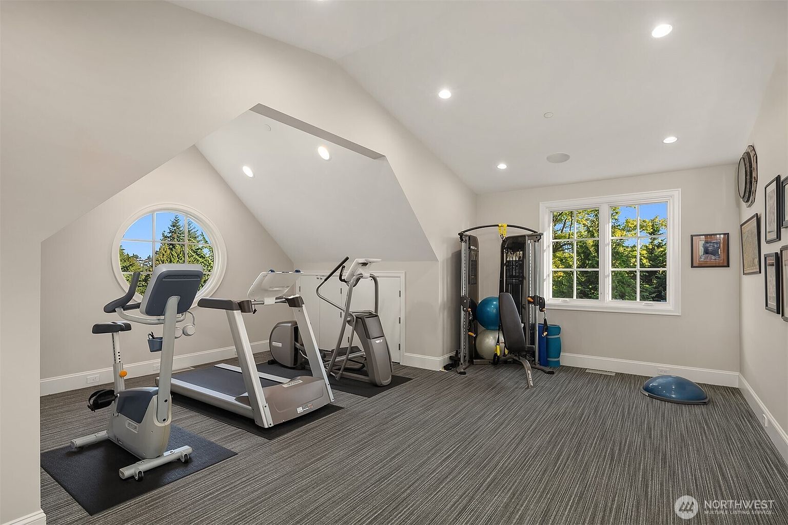 This bright and spacious home gym is situated in an attic-style room with sloped ceilings and recessed lighting. The space is well-equipped with a treadmill, stationary bike, elliptical machine, and a multi-functional cable weight station, all set upon a durable, dark-patterned carpet. Large windows, including a distinctive circular one, provide ample natural light and a pleasant view of the surrounding trees.