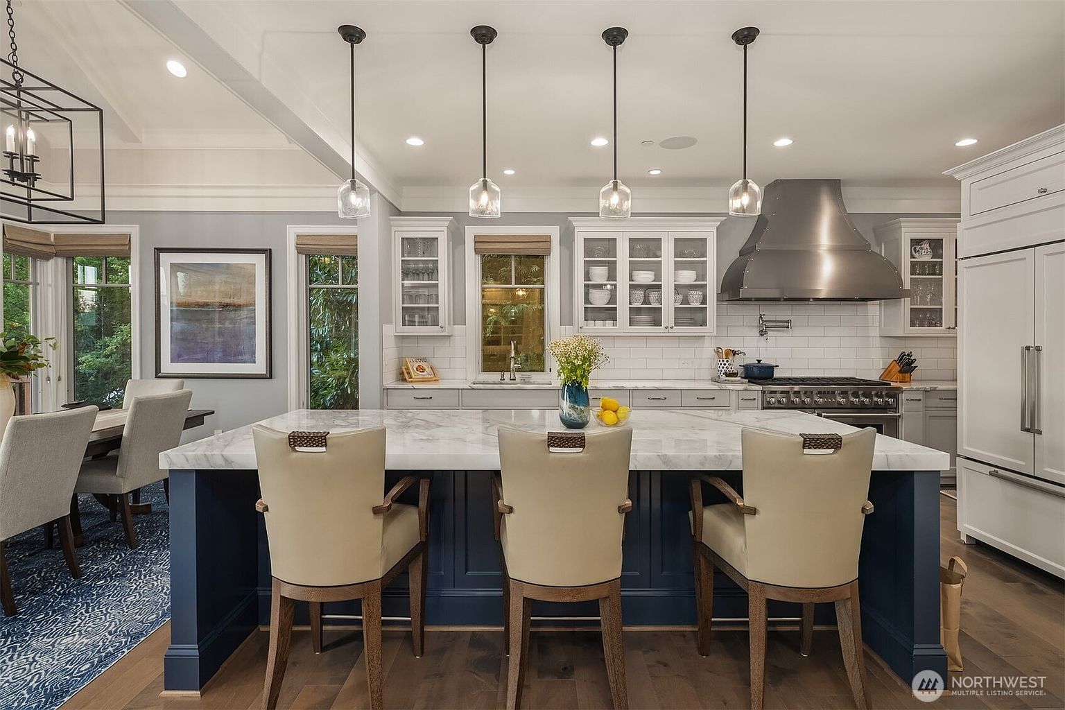 This elegant, high-end kitchen features a large central island with a marble countertop and deep blue cabinetry, complemented by three stylish pendant lights. The space is anchored by a professional-grade stainless steel range hood and white subway tile backsplash, creating a bright and sophisticated atmosphere. A dining area is visible to the left, seamlessly integrating the kitchen into the open-concept living space.