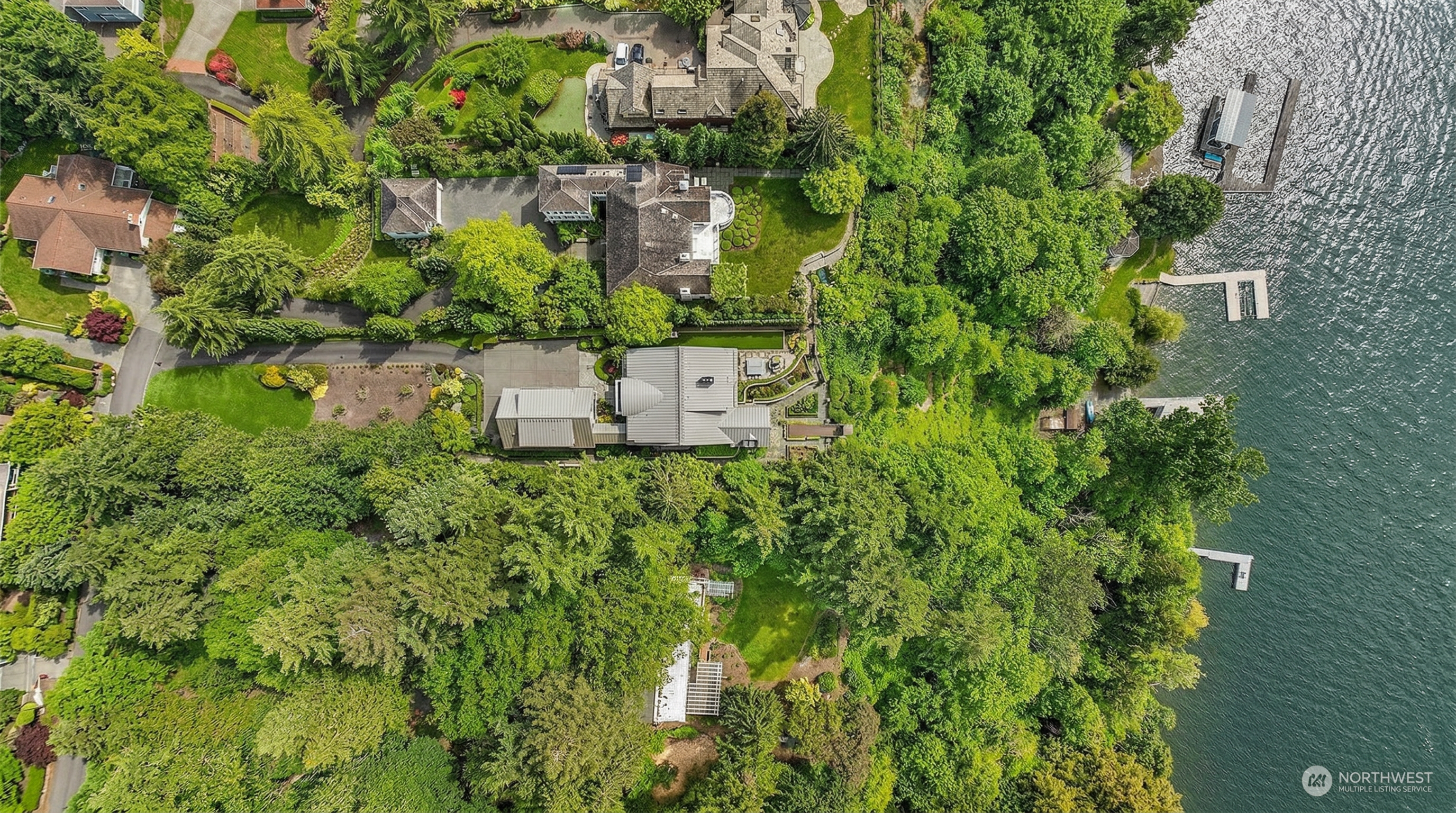 This high-altitude aerial view captures a luxurious waterfront estate nestled within a lush, densely wooded landscape. The property features a modern residence with a distinctive metal roof, expansive manicured lawns, and a private dock extending into the adjacent lake. The perspective highlights the property's secluded, park-like setting and its seamless integration with the natural environment.