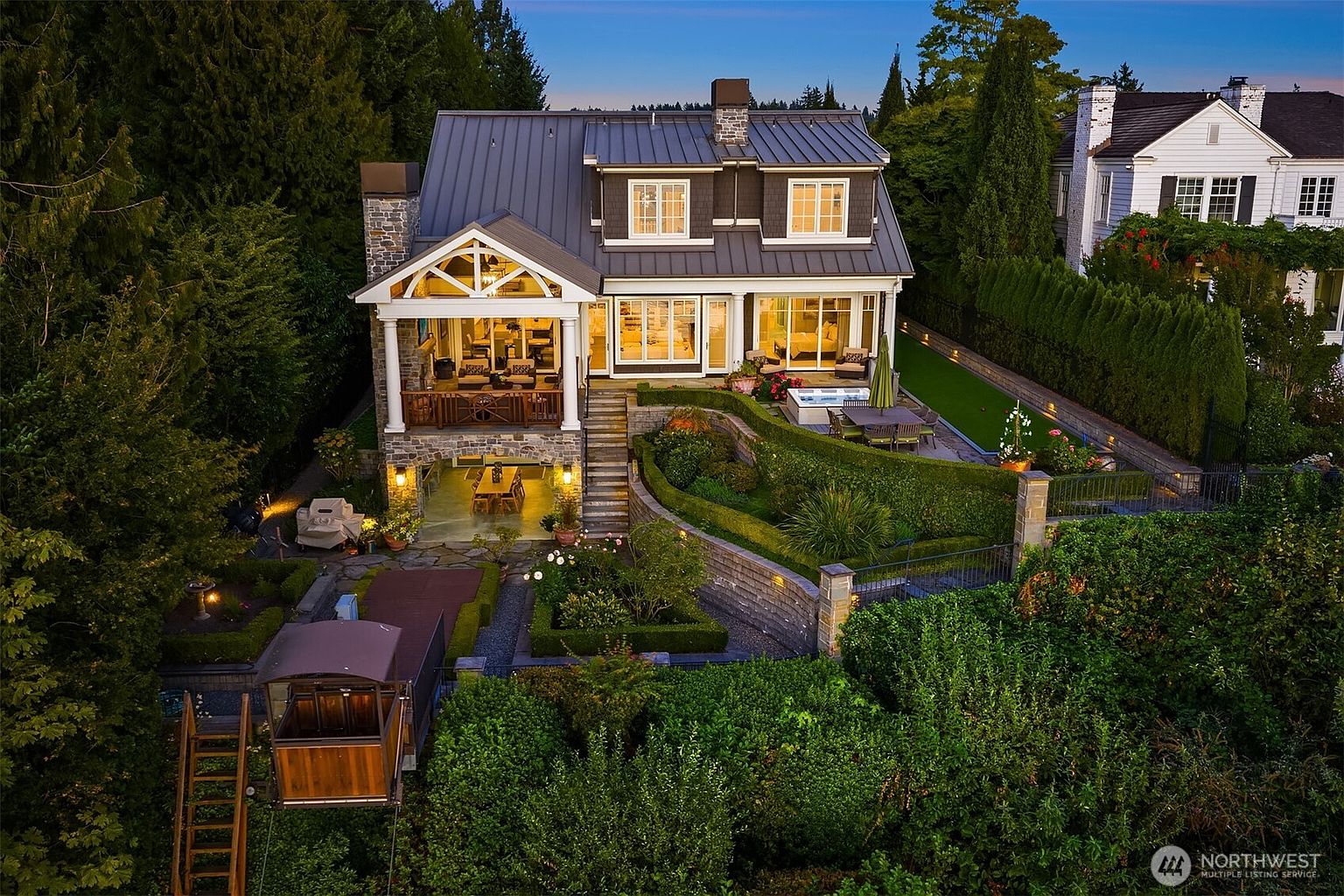 This stunning aerial perspective captures the rear elevation of a luxurious multi-level home at dusk, showcasing a blend of stone and dark metal roofing. The property features a covered upper deck, a walk-out lower level with an outdoor dining area, and a meticulously landscaped terraced garden complete with a hot tub and a private funicular. The warm interior lighting contrasts beautifully with the twilight sky, highlighting the home's sophisticated architectural design and seamless indoor-outdoor living spaces.