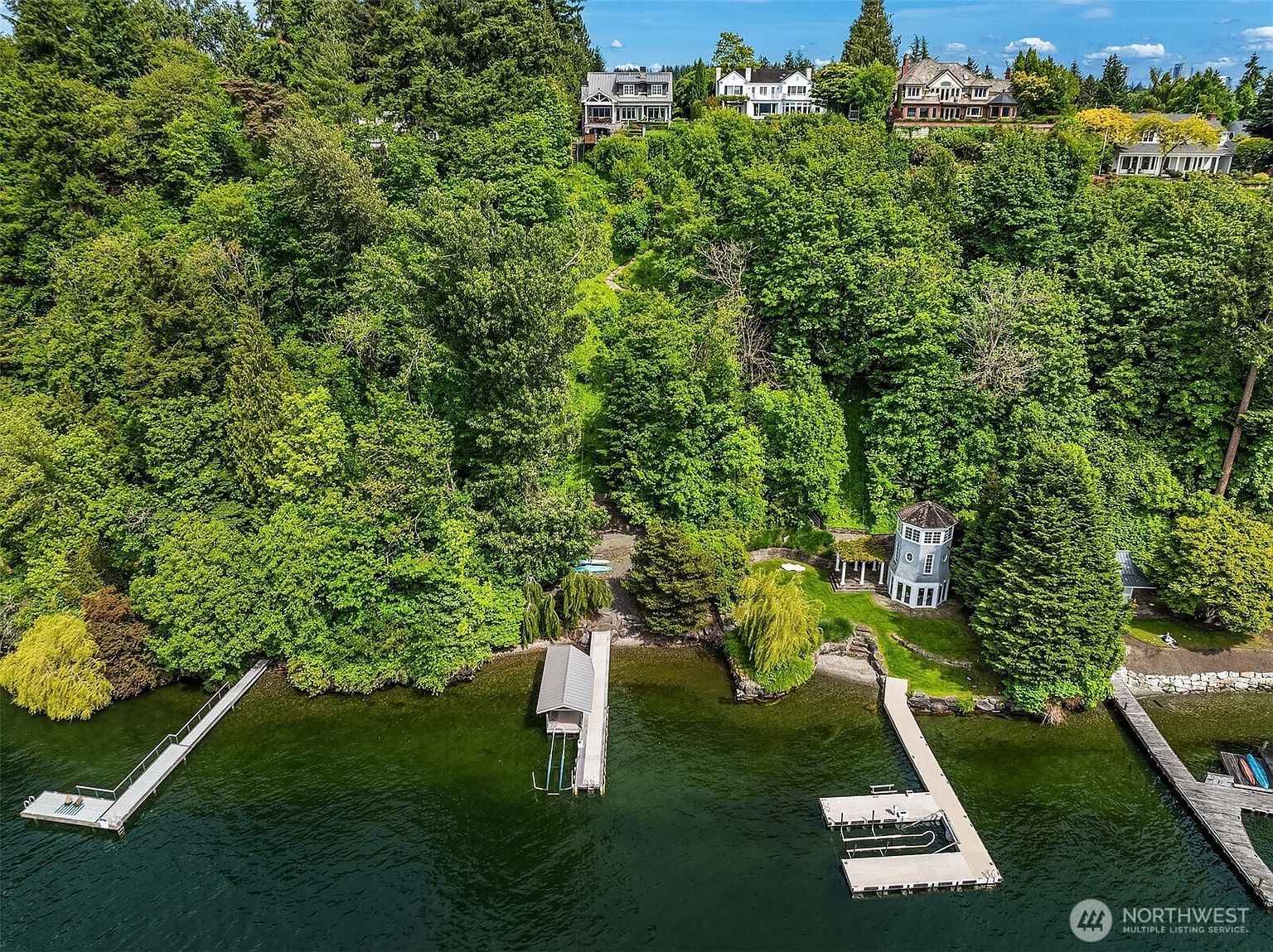 This high-angle aerial view captures a lush, wooded waterfront property featuring a private dock and a unique, multi-story turret-style structure near the shoreline. The dense forest canopy leads up a steep slope to several large, upscale residences perched at the top, offering a secluded and serene lakeside atmosphere. The perspective emphasizes the expansive natural landscape and the direct water access, highlighting the property's exclusive and tranquil setting.