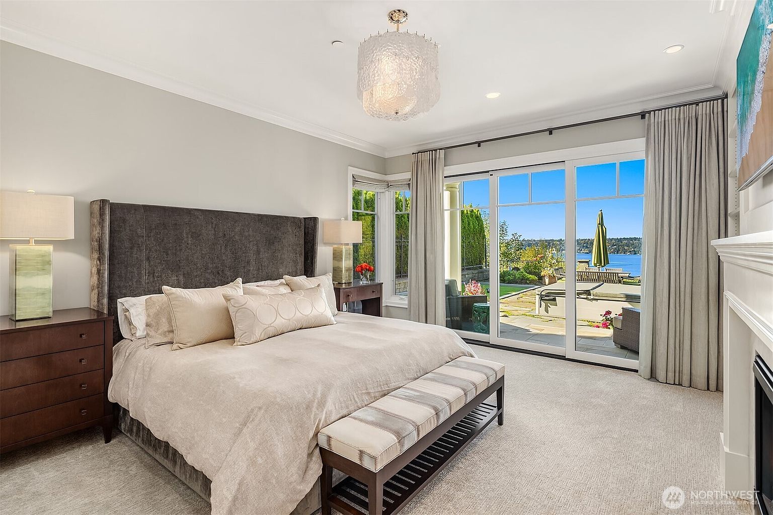 This elegant primary bedroom features a plush, dark velvet upholstered headboard and a neutral-toned bedding ensemble, creating a sophisticated and serene atmosphere. Large sliding glass doors provide an expansive view of a private patio and a scenic waterfront, seamlessly blending indoor comfort with outdoor beauty. The room is accented by a unique, textured chandelier and matching bedside lamps, enhancing the luxurious and tranquil aesthetic.