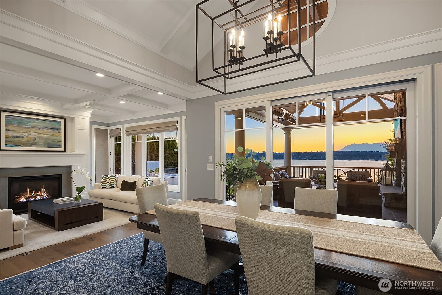 This elegant open-concept living and dining space features a sophisticated blend of modern and traditional design, highlighted by a large dining table under a striking geometric chandelier. The room flows seamlessly into a cozy living area with a fireplace and expansive sliding glass doors that frame a breathtaking sunset view over the water. The neutral color palette, coffered ceilings, and high-end finishes create a luxurious and inviting atmosphere perfect for entertaining.