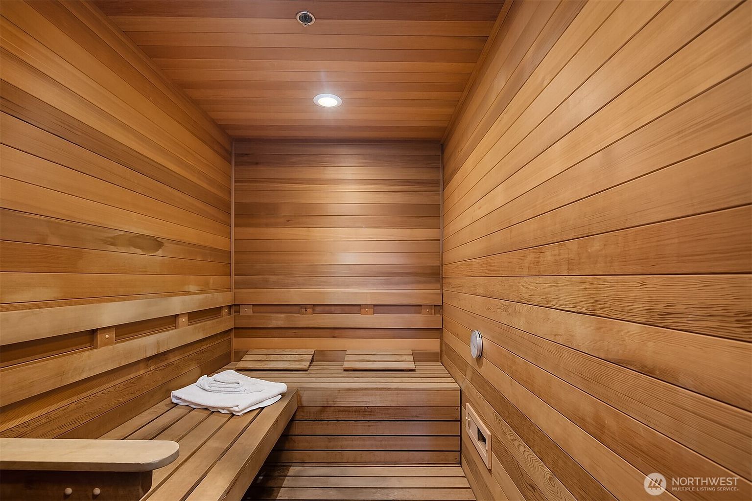 This image showcases a compact, custom-built cedar sauna, characterized by its warm, natural wood paneling and tiered bench seating. The space is illuminated by a recessed ceiling light, creating a cozy and inviting atmosphere for relaxation. A neatly folded white towel rests on the lower bench, emphasizing the spa-like quality of this home amenity.