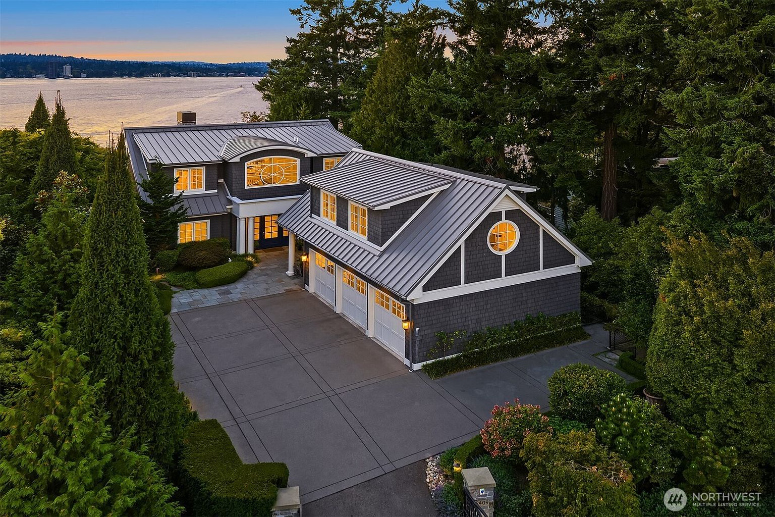 This high-angle aerial shot captures a stunning, dark-shingled waterfront residence at dusk, highlighting its sophisticated architectural design and metal roofing. The property features a spacious three-car garage, a stone-paved driveway, and lush, mature landscaping that frames the home against the serene backdrop of a large lake. The warm glow from the interior windows creates an inviting atmosphere, emphasizing the home's luxury and prime location.