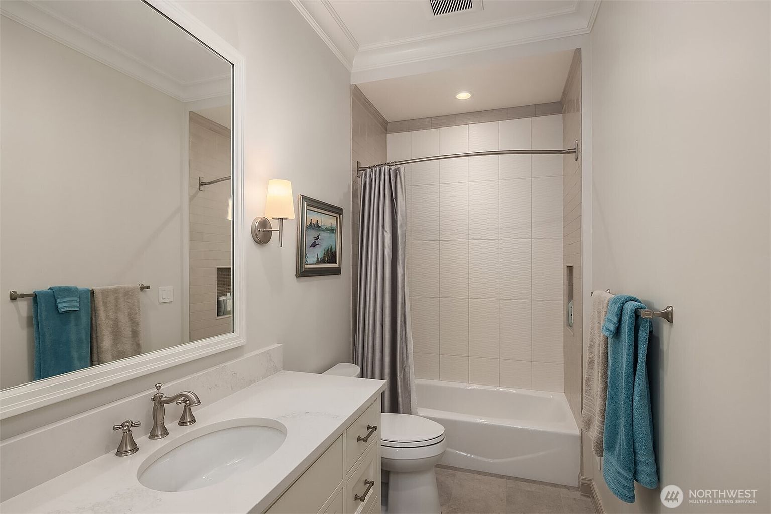 This clean and well-lit bathroom features a white vanity with a marble countertop, a large framed mirror, and a shower-tub combination with textured white tile walls. The space is accented with soft blue towels and a framed piece of wall art, creating a bright and inviting atmosphere. The perspective is a straight-on shot capturing the functional layout of the room.