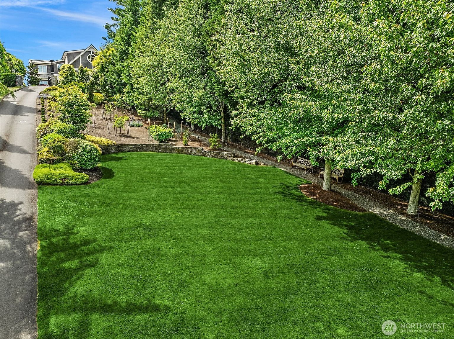 This image captures a lush, expansive backyard featuring a meticulously manicured green lawn that slopes gently upward toward a wooded area. A stone retaining wall separates the lawn from a landscaped hillside, while a paved driveway leads up to a multi-story home in the background. The scene is bathed in bright, natural sunlight, creating a serene and inviting outdoor living space.