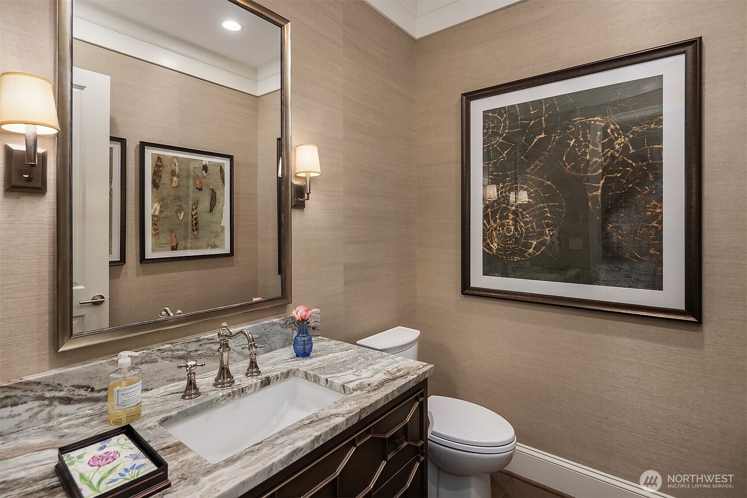 This elegant powder room features sophisticated textured wallpaper in a warm neutral tone, complemented by a marble-topped vanity with a dark wood base. A large, framed mirror reflects the space, which is accented by tasteful wall sconces and framed artwork, creating a refined and welcoming atmosphere. The clean lines and high-end finishes suggest a luxurious and well-appointed home.