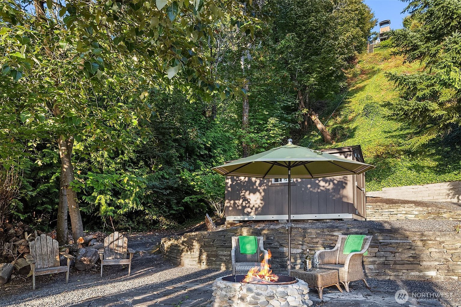 This inviting outdoor patio area features a stone fire pit as the centerpiece, surrounded by a mix of rustic wooden Adirondack chairs and comfortable wicker seating with green cushions. A large green umbrella provides shade, while a stone retaining wall and a small shed nestled against a lush, wooded hillside create a cozy, secluded atmosphere. The scene is captured from a ground-level perspective, emphasizing the tranquil and private nature of this backyard retreat.